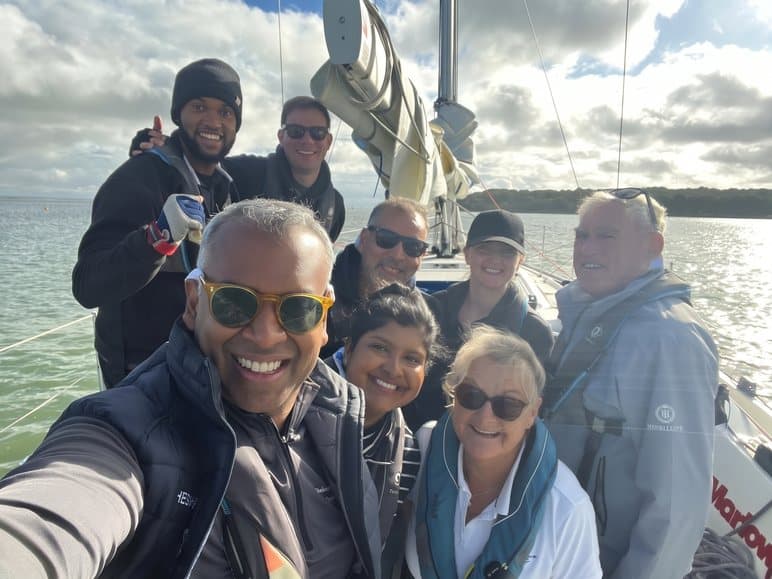A crew selfie taken mid-channel, with the mainsail and a cloudy sky behind the group.